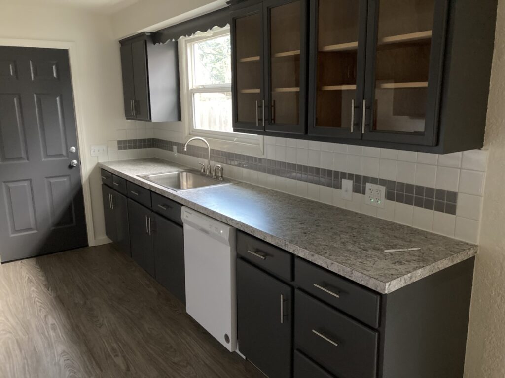 Kitchen remodel in Veneta, Oregon featuring dark gray cabinets, stone-look countertops, tile backsplash, stainless steel sink, and wood-look flooring.